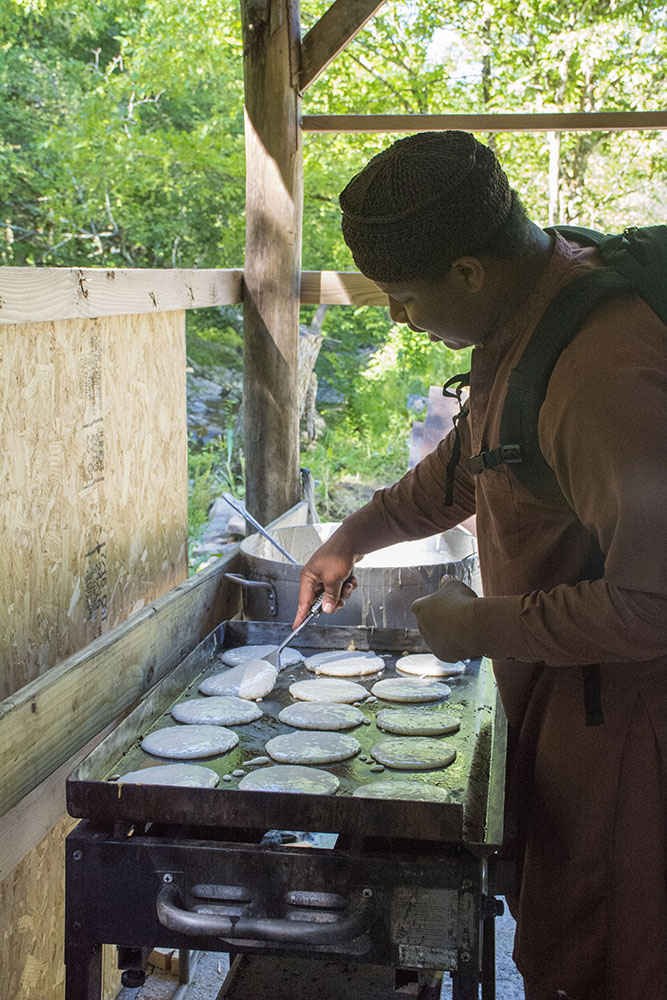 A camp cook flipping pancakes on an outdoor griddle beside the woods